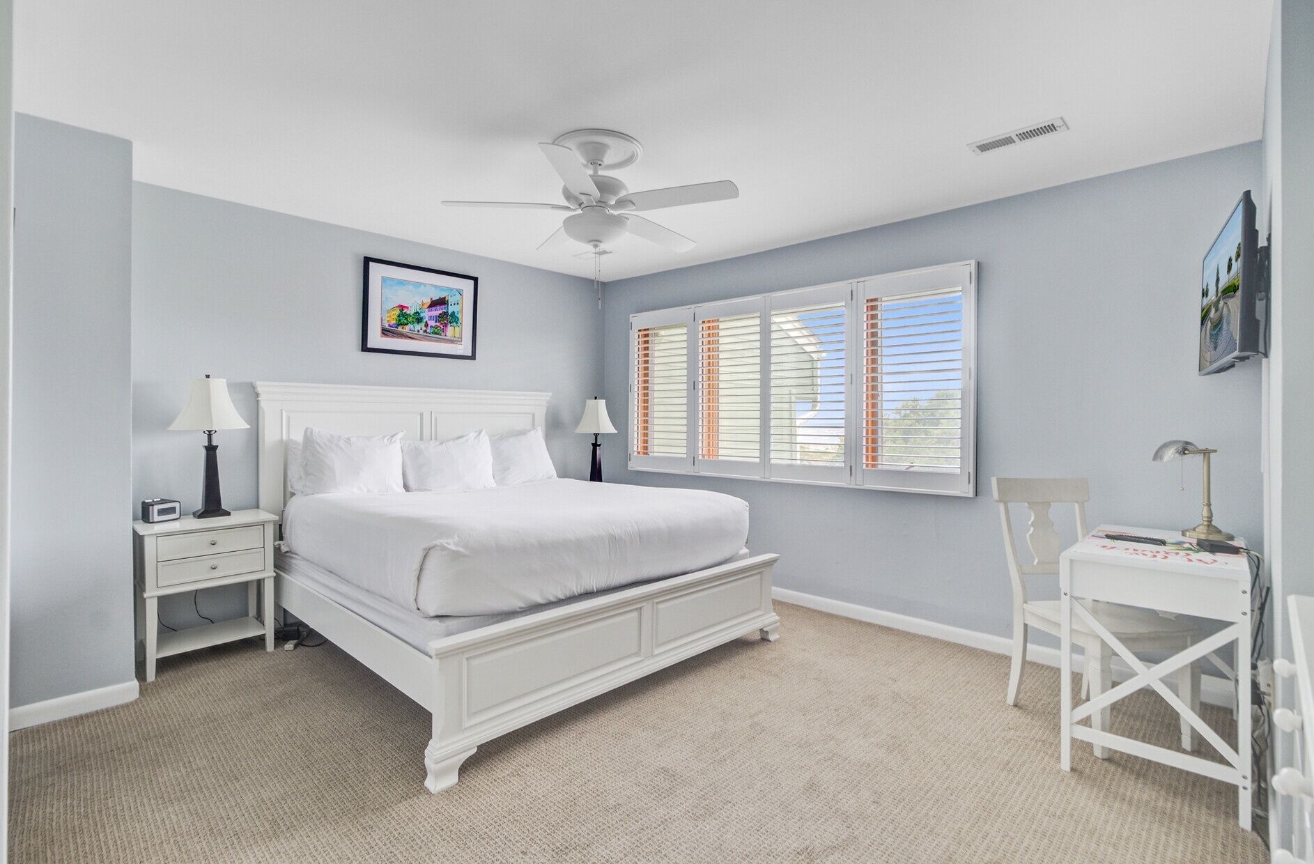 A bedroom with a white bed, two bedside tables, a ceiling fan, a window with blinds, and a framed picture on the wall.