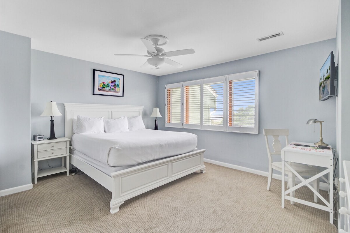 A bedroom with a white bed, two bedside tables, a ceiling fan, a window with blinds, and a framed picture on the wall.