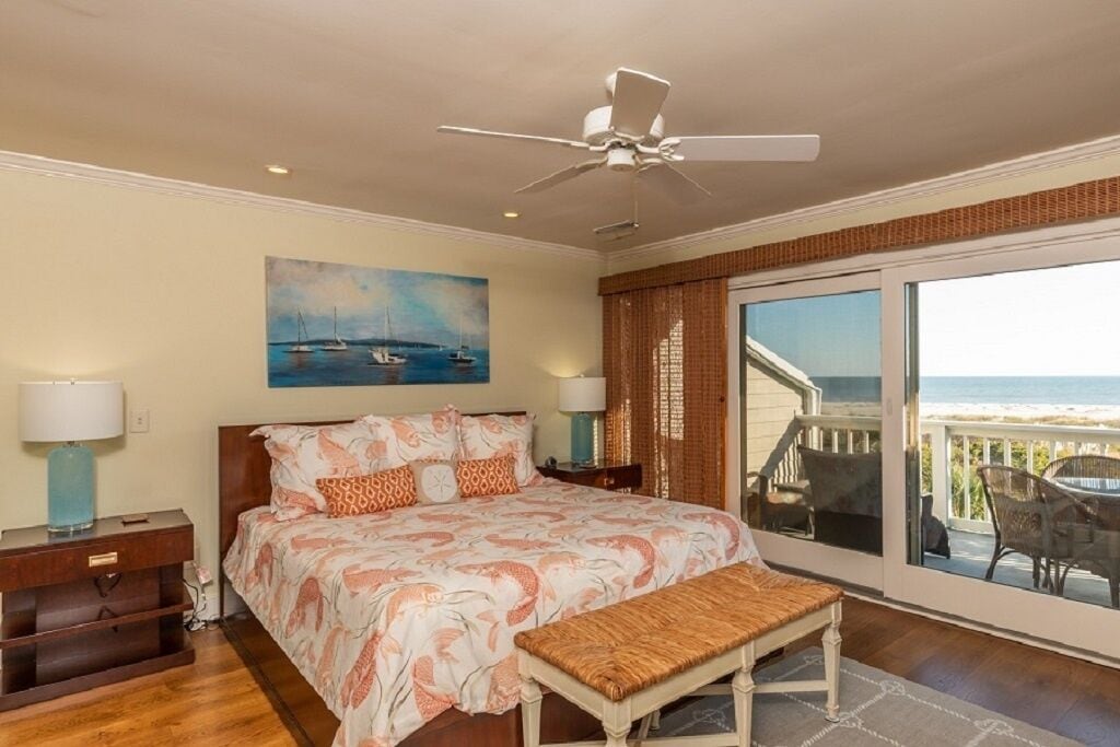 A bedroom with a bed, a ceiling fan, a painting of boats, a nightstand, a bench, and a view of the beach through a sliding door.