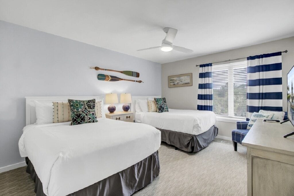 A room with two beds, a ceiling fan, a window with striped curtains, and a desk with a mirror.