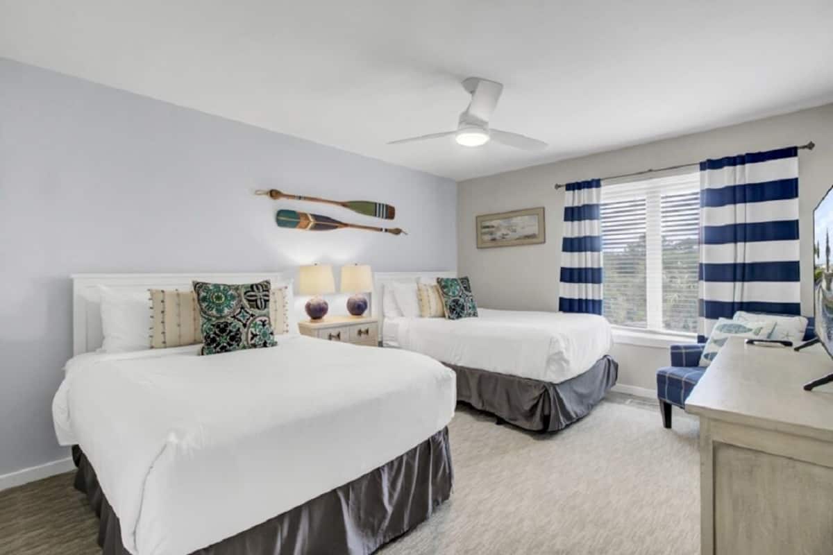 Una habitación con dos camas, un ventilador de techo, una ventana con cortinas a rayas y un escritorio con espejo.