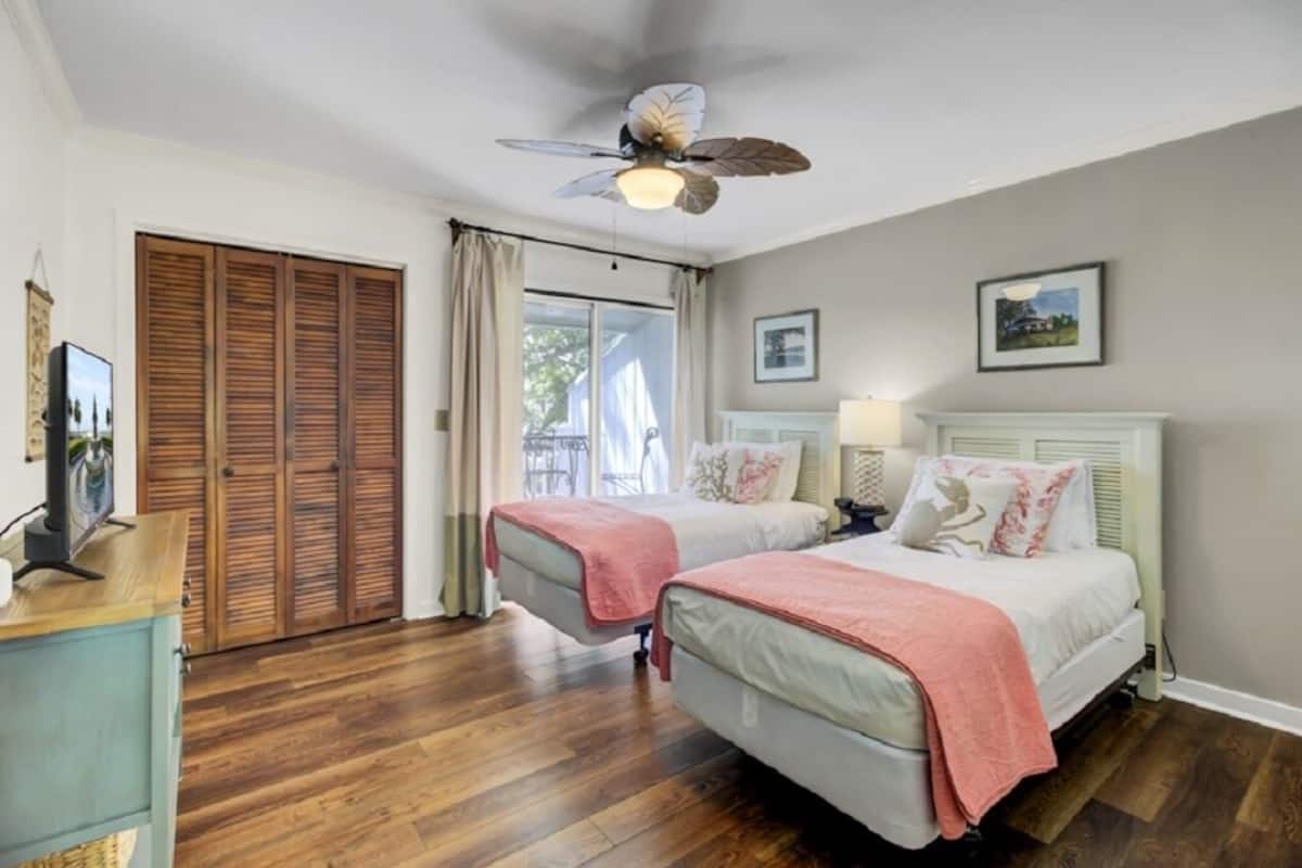 Un dormitorio con dos camas, un ventilador de techo, una ventana con cortinas, suelo de madera y un aparador blanco.