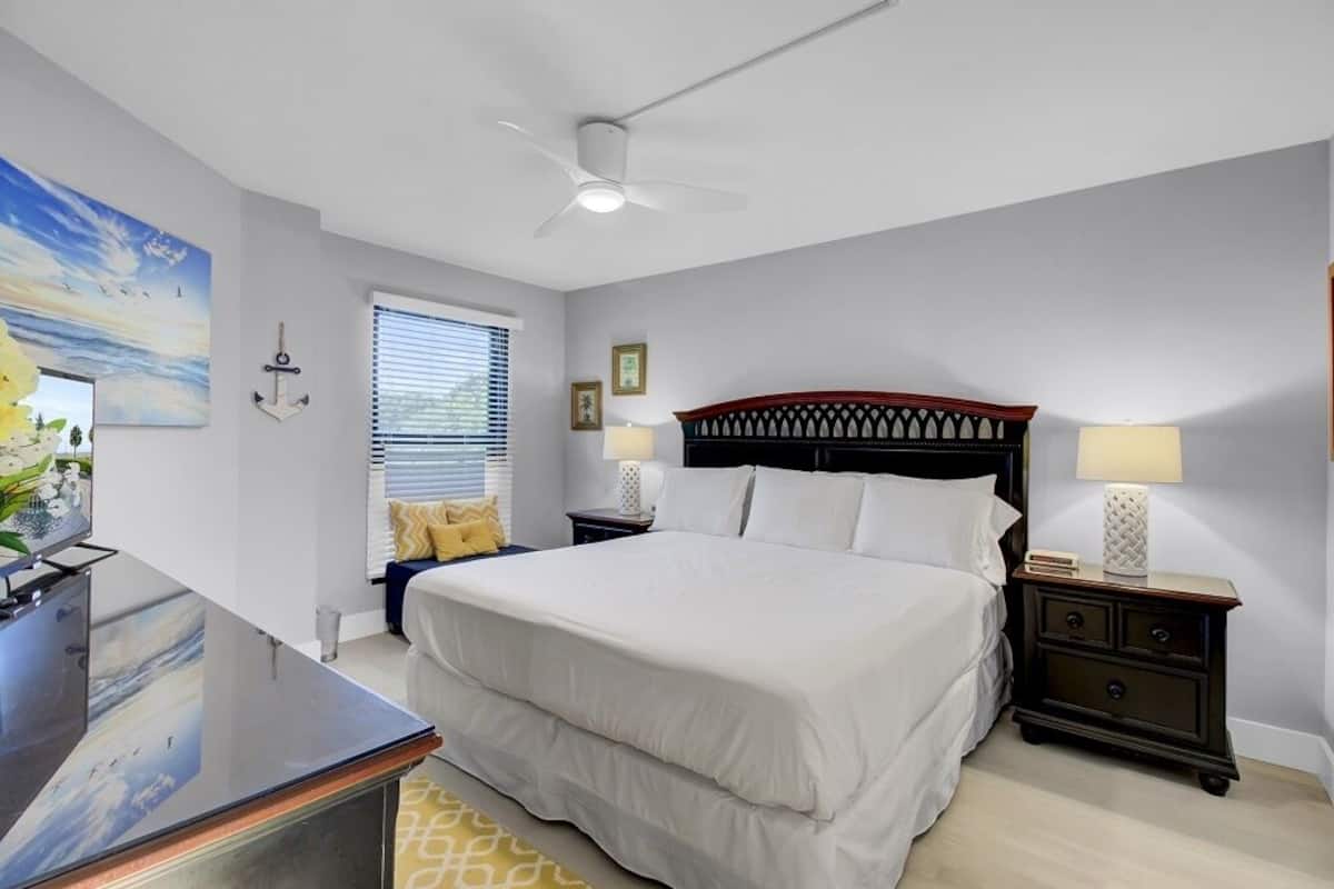 Un dormitorio con cama, mesitas de noche, escritorio, ventilador de techo y ventana con persianas.