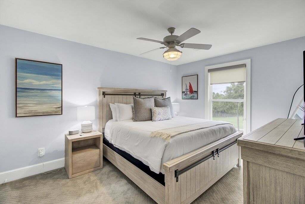 A bedroom with a bed, bedside table, nightstand, ceiling fan, and a window with a view.