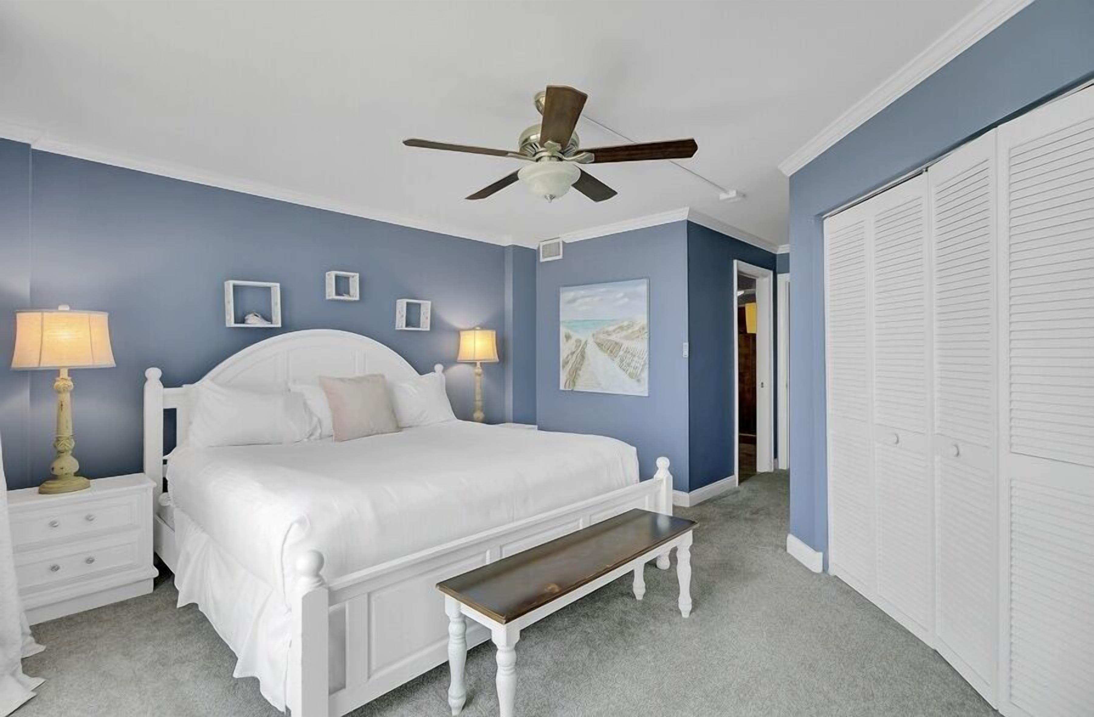A bedroom with a bed, bedside tables, a ceiling fan, a nightstand, a bench, and a closet.