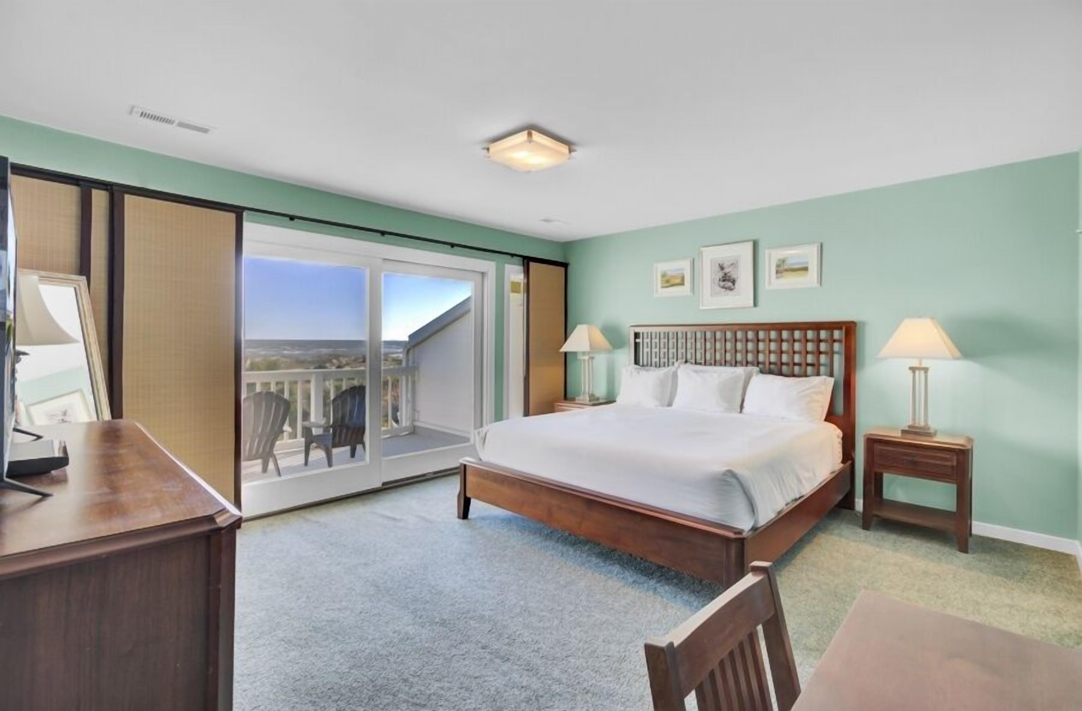 A bedroom with a bed, two bedside tables, a desk, a chair, and a view through a sliding door to an outdoor area.