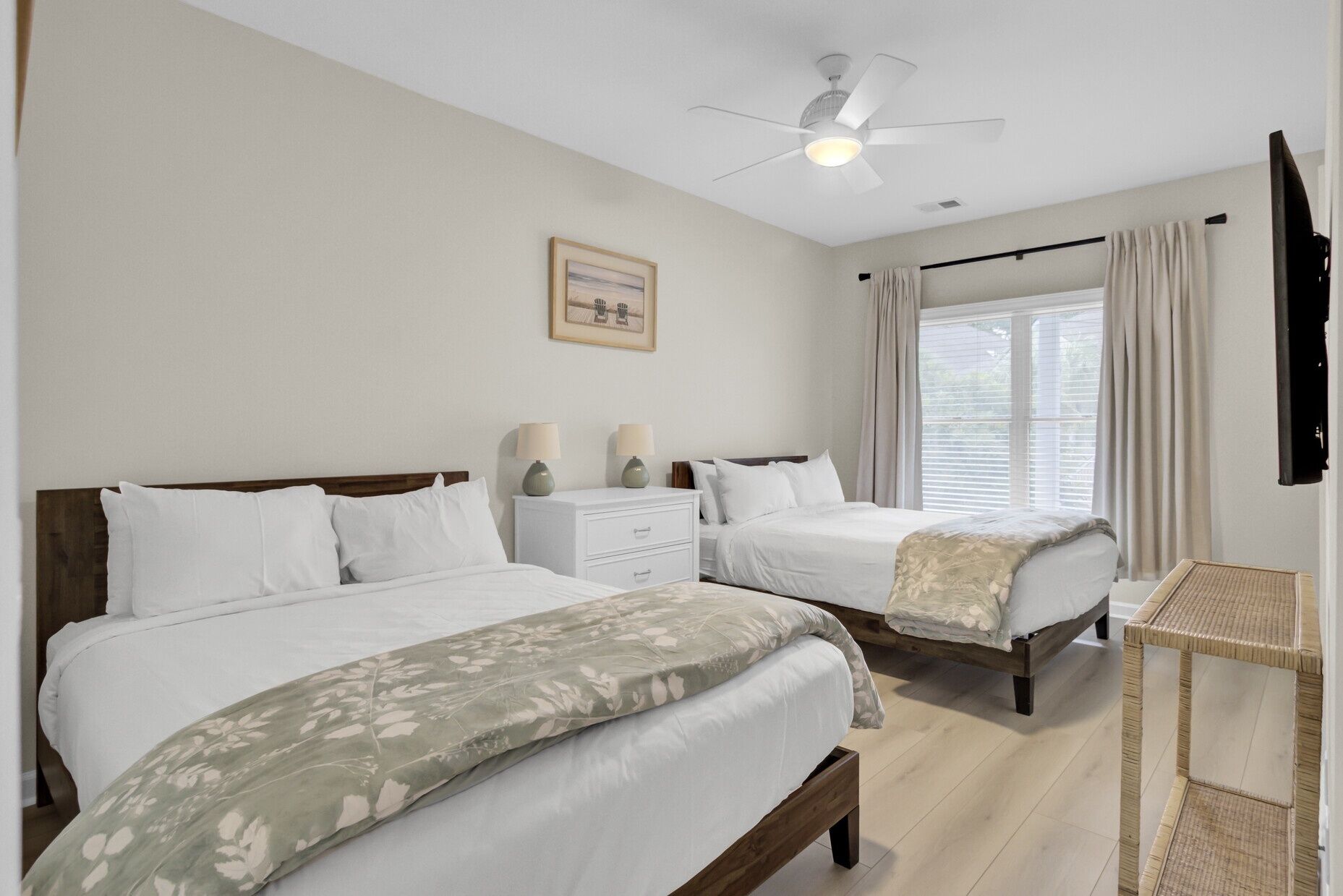 A hotel room with two beds, a ceiling fan, a window with curtains, a bedside table with lamps, and a framed picture on the wall.