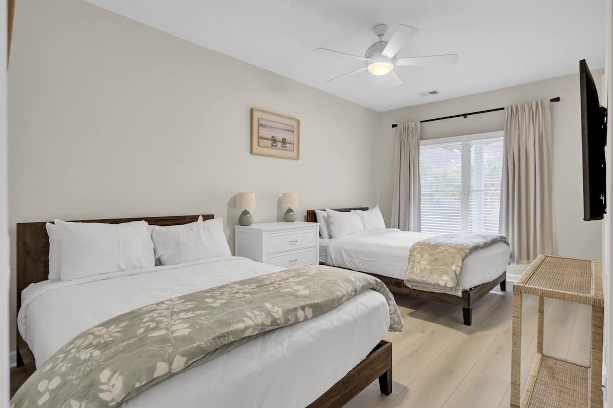 Habitación de hotel con dos camas, un ventilador de techo, una ventana con cortinas, una mesita de noche con lámparas y un cuadro enmarcado en la pared.