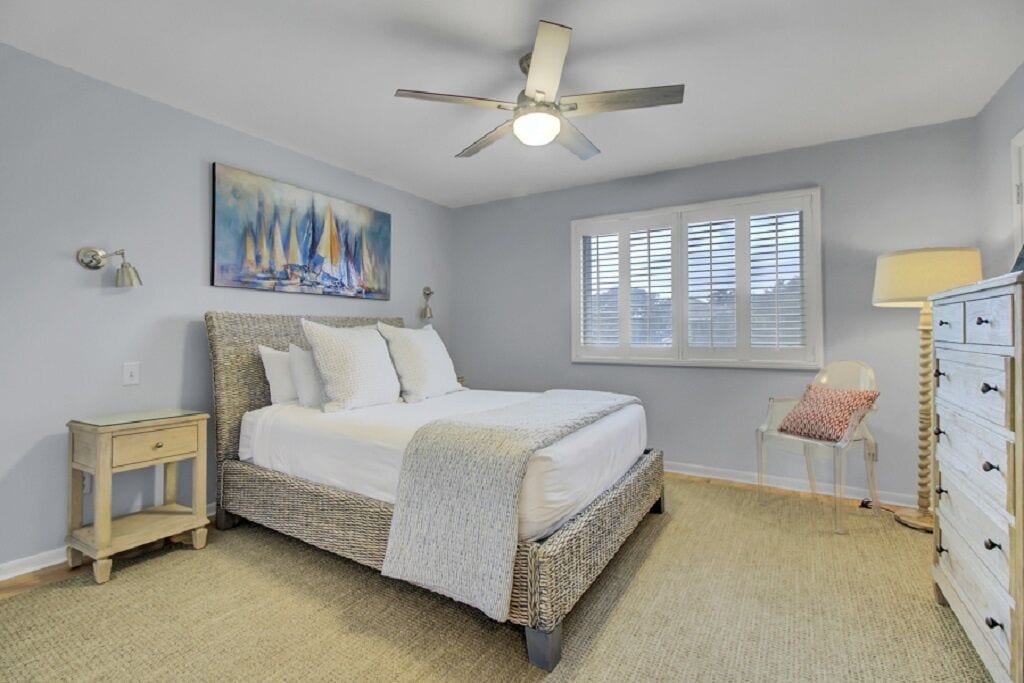 A bedroom with a bed, a ceiling fan, a window with blinds, a nightstand, a dresser, and a painting on the wall.