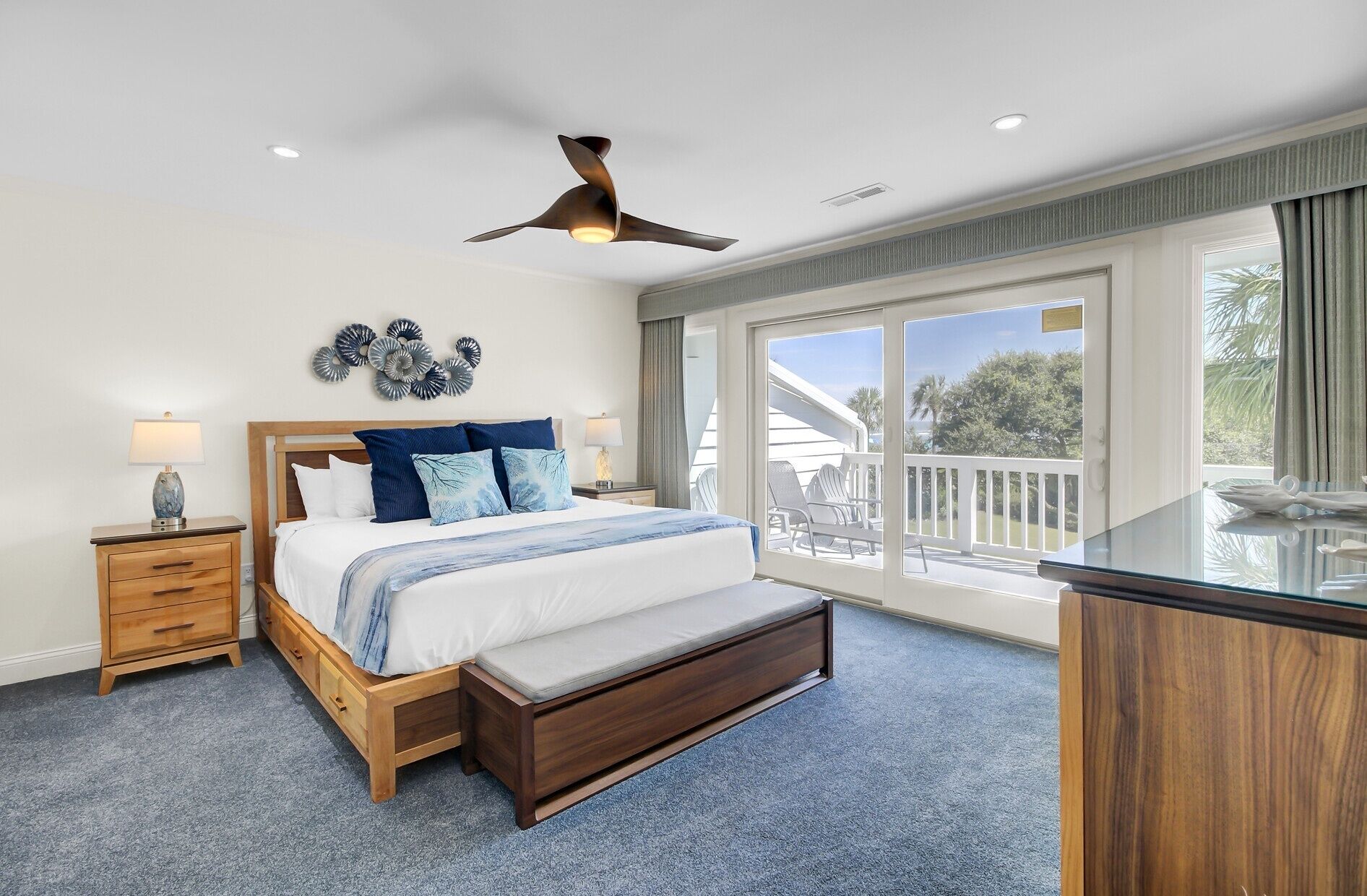 A bedroom with a bed, bedside tables, a bench, a ceiling fan, and a balcony with outdoor furniture.
