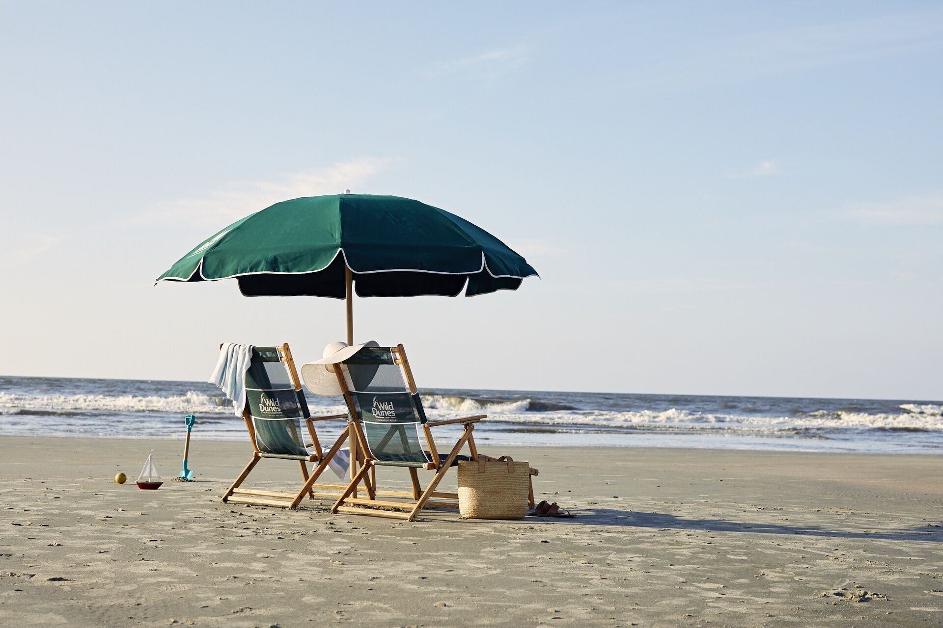 A beach with two beach chairs under a green umbrella, a small boat, and a beach bag on the sand.