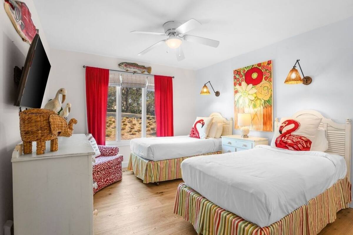 Un dormitorio con dos camas, ventilador de techo, televisor, ventana con cortinas rojas y un cuadro en la pared.