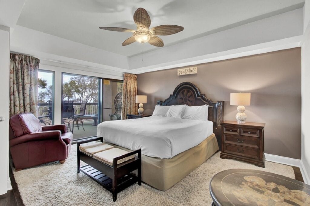 A bedroom with a bed, a desk, a chair, a ceiling fan, and a view of the outdoors.