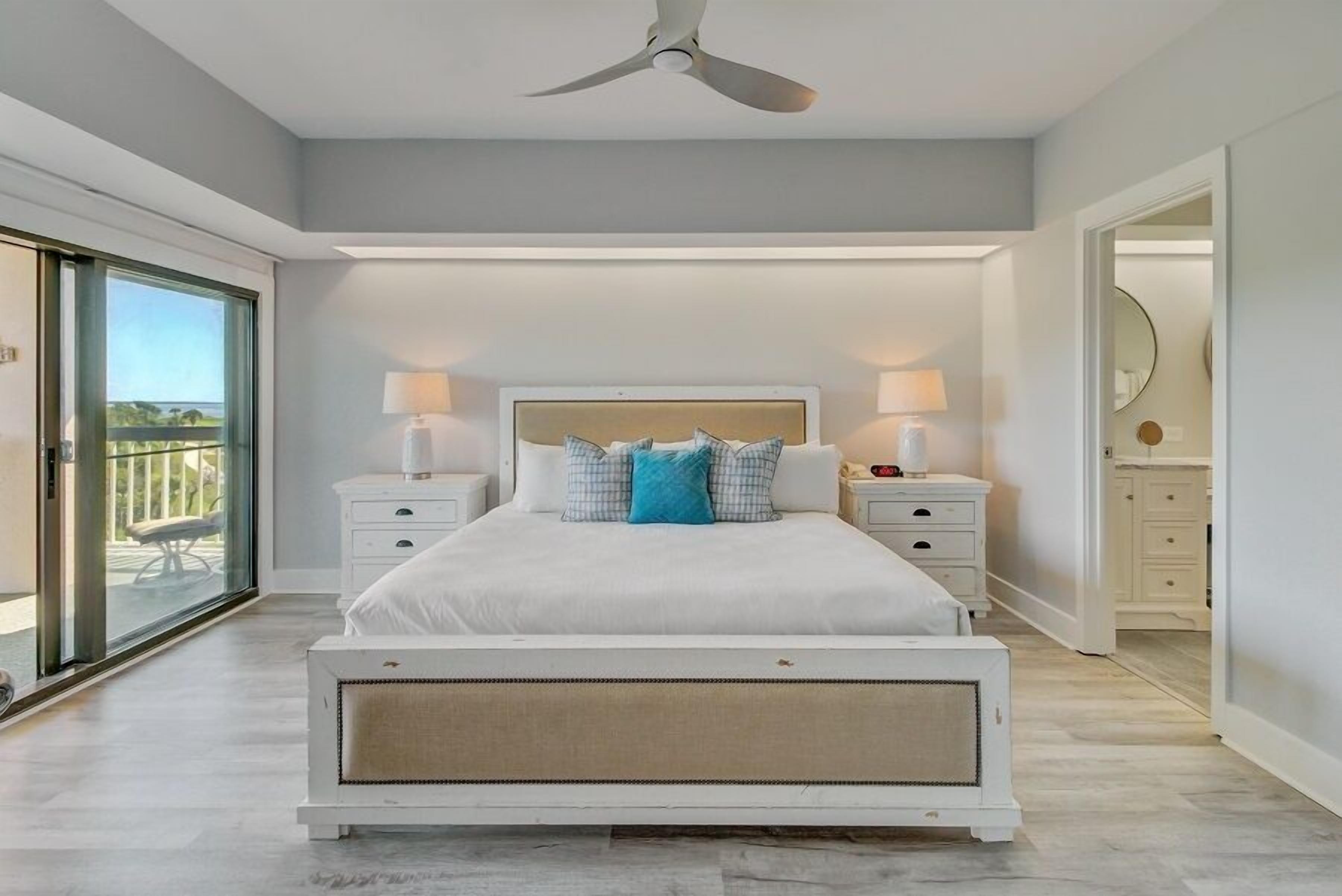 A modern bedroom with a large bed, two bedside tables, a ceiling fan, and a sliding glass door leading to a balcony.