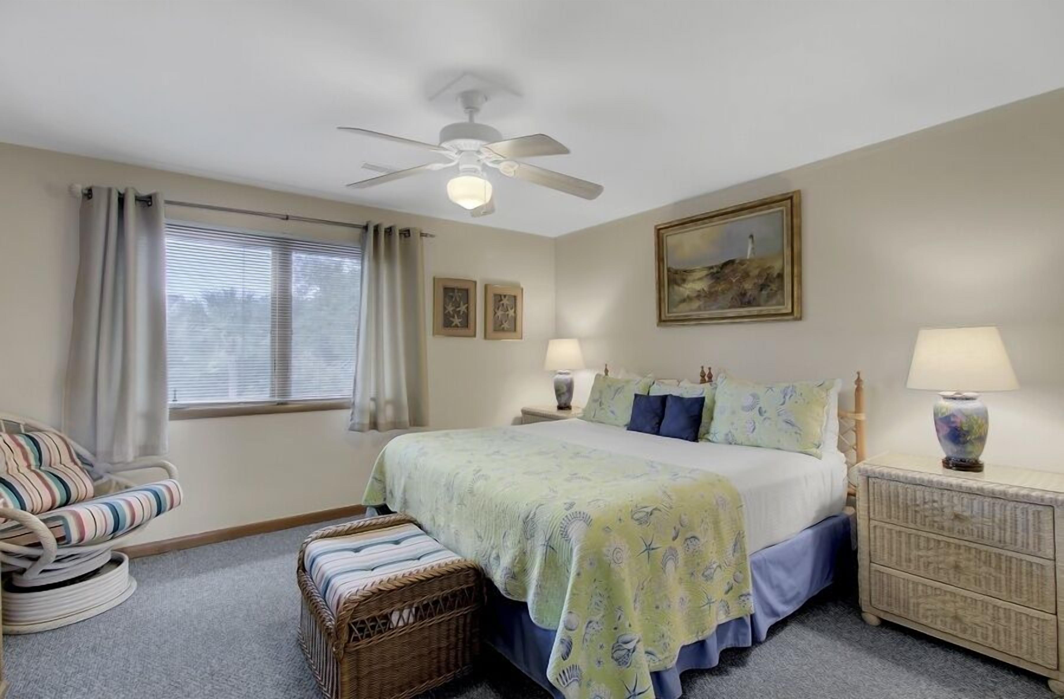 A bedroom with a bed, a chair, a nightstand, a lamp, a ceiling fan, and a window with curtains.