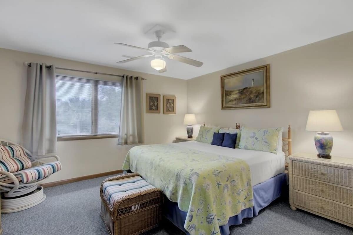 Un dormitorio con cama, una silla, mesita de noche, lámpara, ventilador de techo y ventana con cortinas.