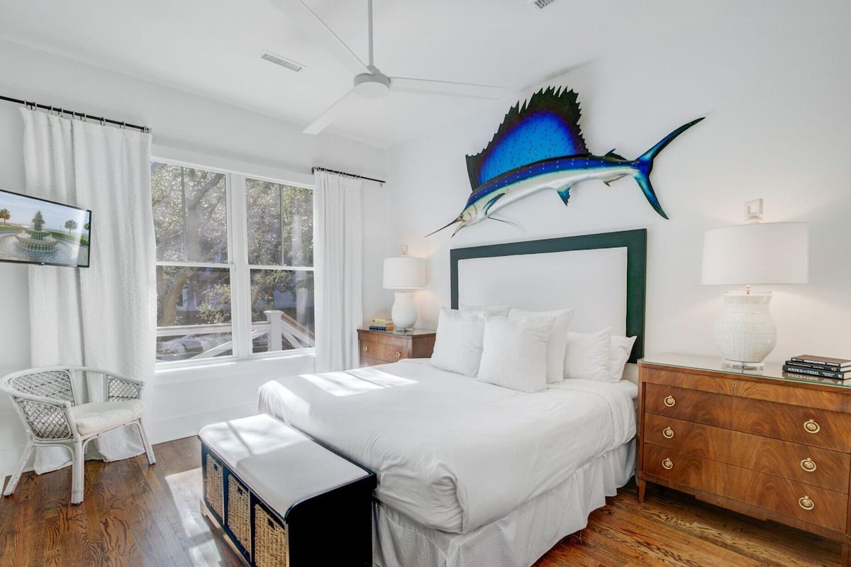 A bedroom with a bed, a nightstand, a chair, a window, and a wall-mounted artwork of a blue fish.