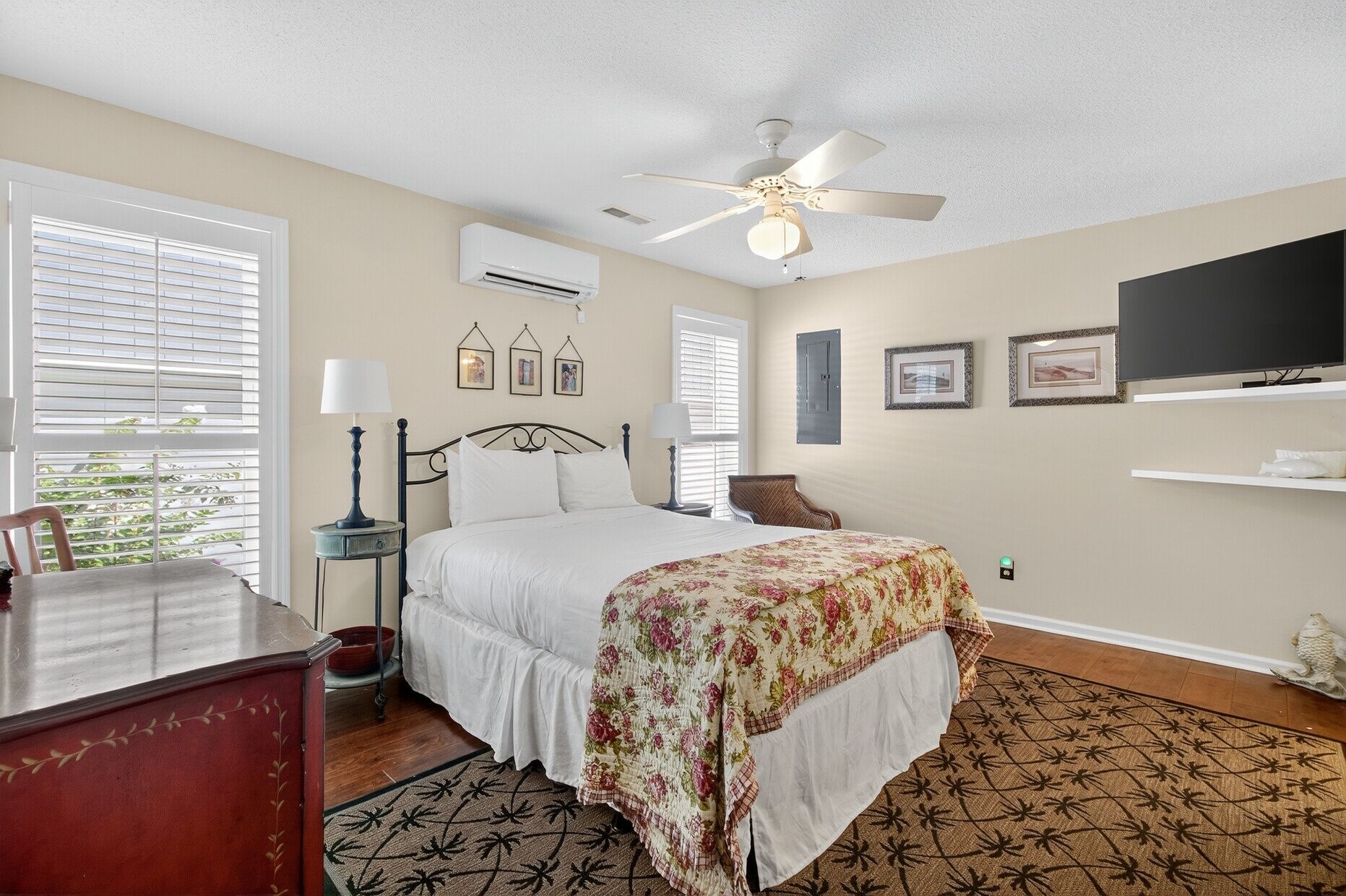 A bedroom with a bed, a ceiling fan, an air conditioner, a TV, framed pictures on the wall, and a window with blinds.