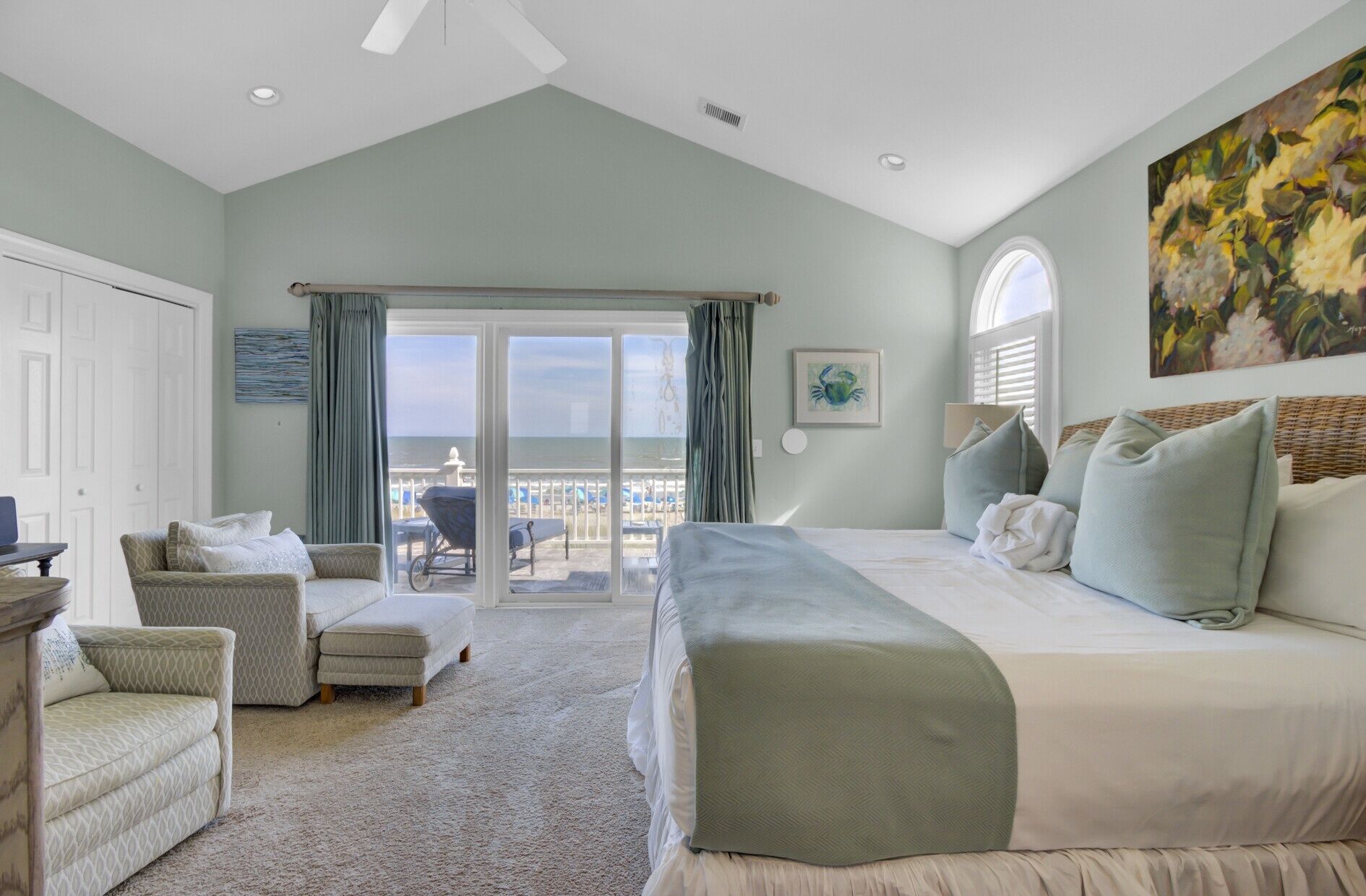 A bedroom with a large bed, a sofa, a chair, and a view of the ocean through a sliding door.