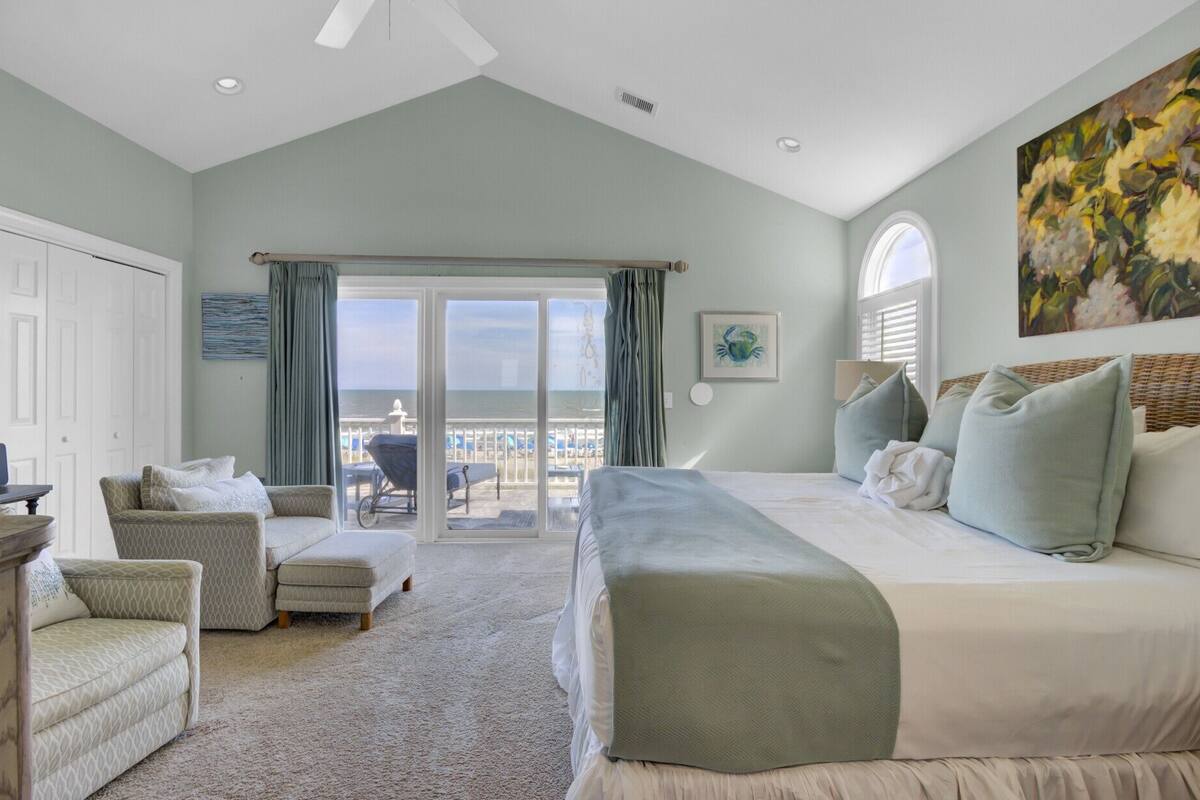 A bedroom with a large bed, a sofa, a chair, and a view of the ocean through a sliding door.