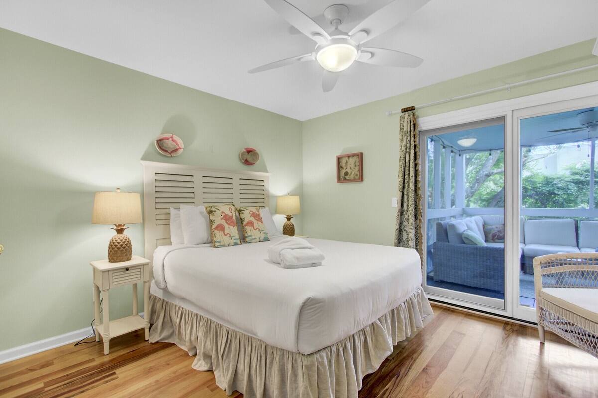 A bedroom with a bed, nightstands, a ceiling fan, a view to the outside, and a balcony with wicker furniture.