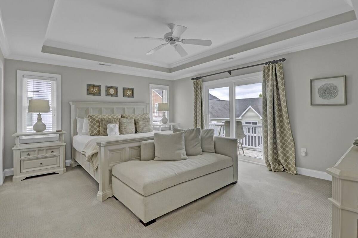 A bedroom with a bed, a white sofa, a nightstand, a ceiling fan, and a window with curtains.
