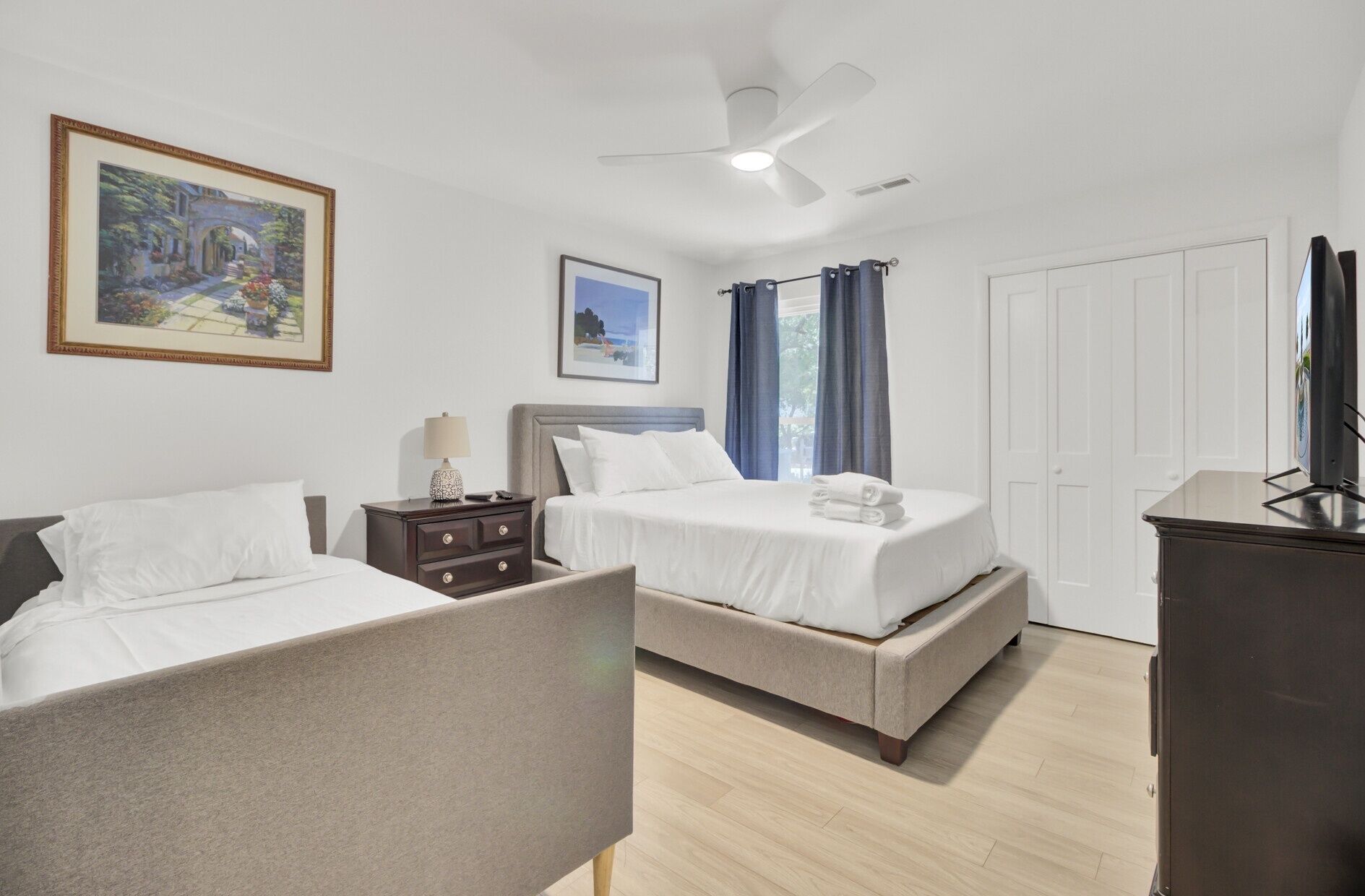 A hotel room with two beds, a ceiling fan, a nightstand, a lamp, and a painting on the wall.