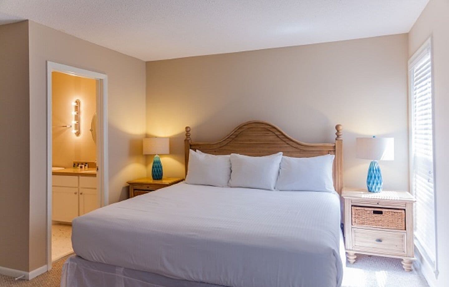 A bedroom with a large bed, two bedside tables with lamps, a wooden headboard, and a door leading to a bathroom.