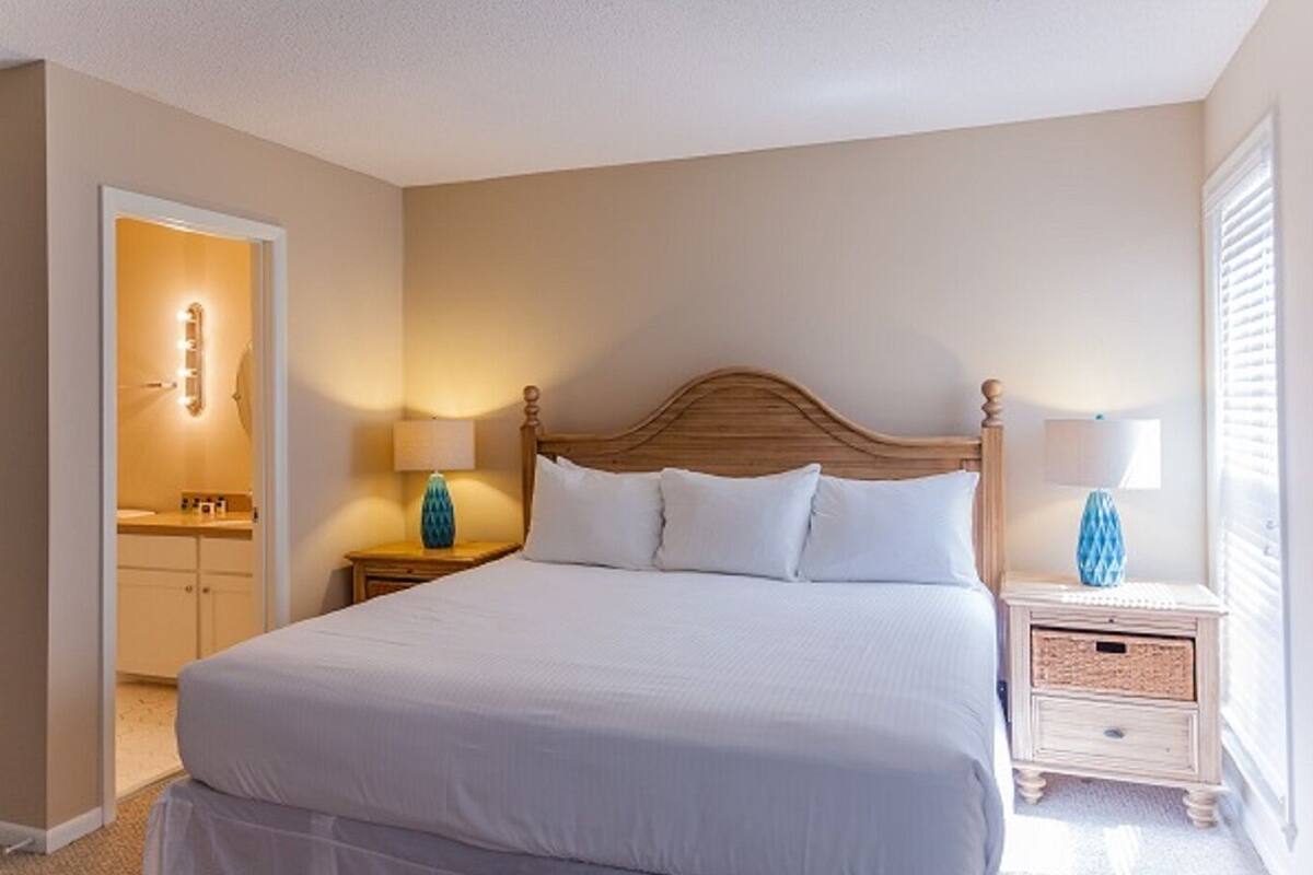 A bedroom with a large bed, two bedside tables with lamps, a wooden headboard, and a door leading to a bathroom.