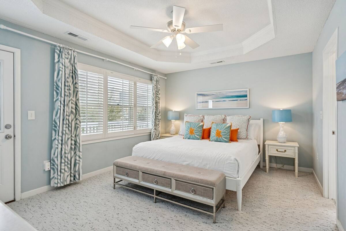 A bedroom with a bed, bedside tables, a bench, a ceiling fan, and a window with blinds.