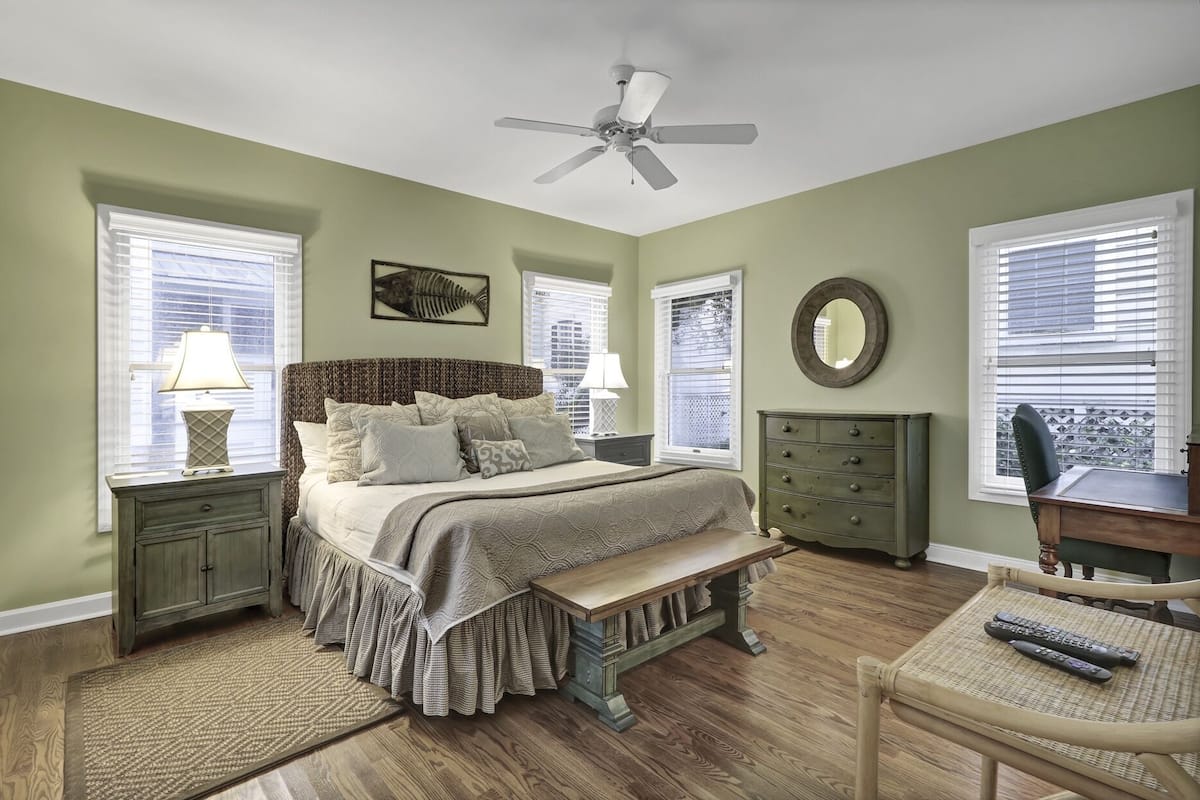 A bedroom with a bed, bedside tables, a dresser, a chair, a ceiling fan, and windows with blinds.
