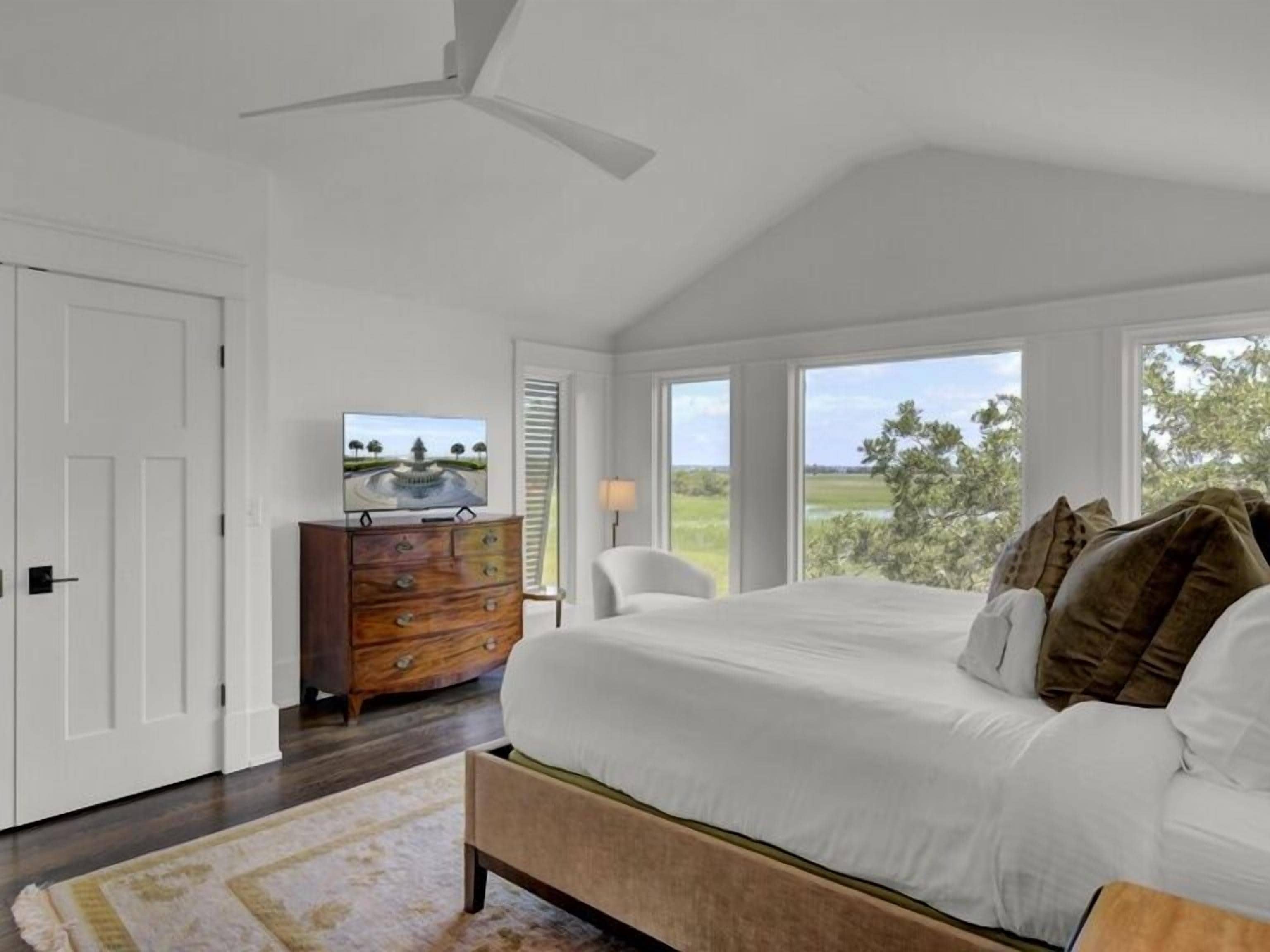 A bedroom with a large bed, a dresser, a television, and a view of the outdoors.