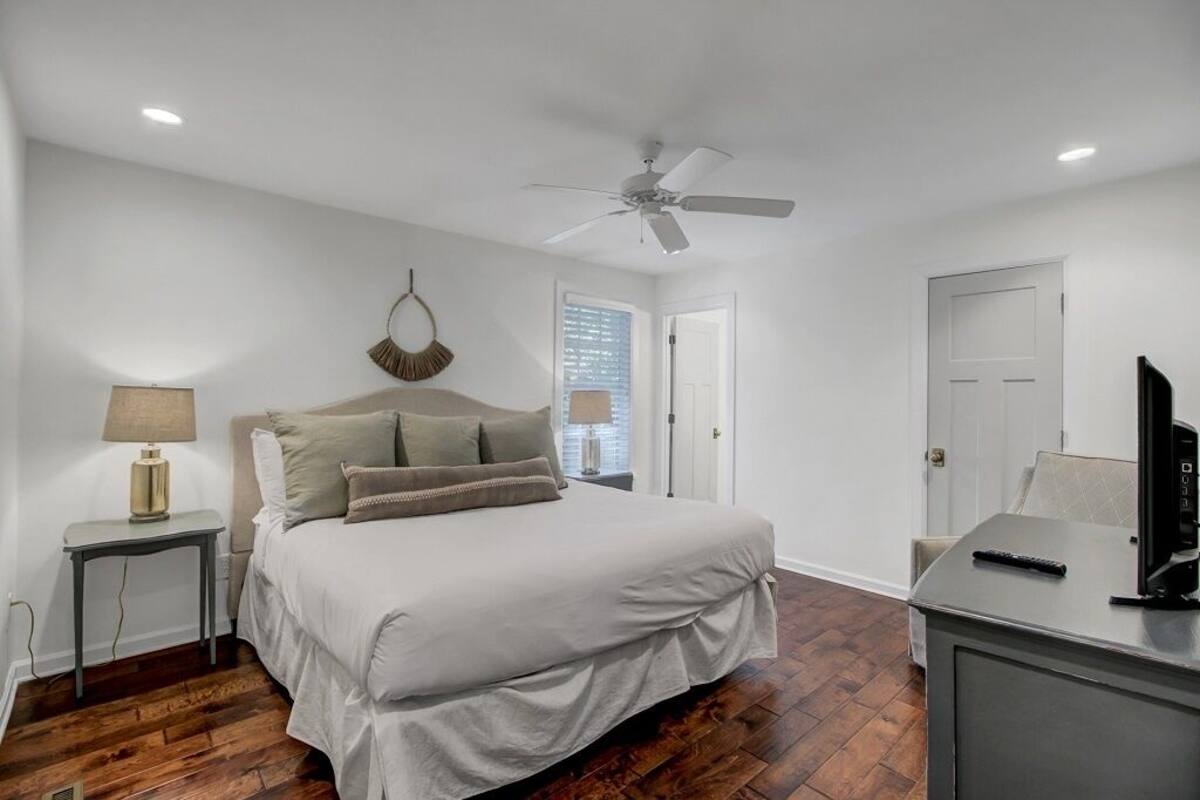 A bedroom with a bed, a ceiling fan, a nightstand with a lamp, a desk with a TV, and a door.