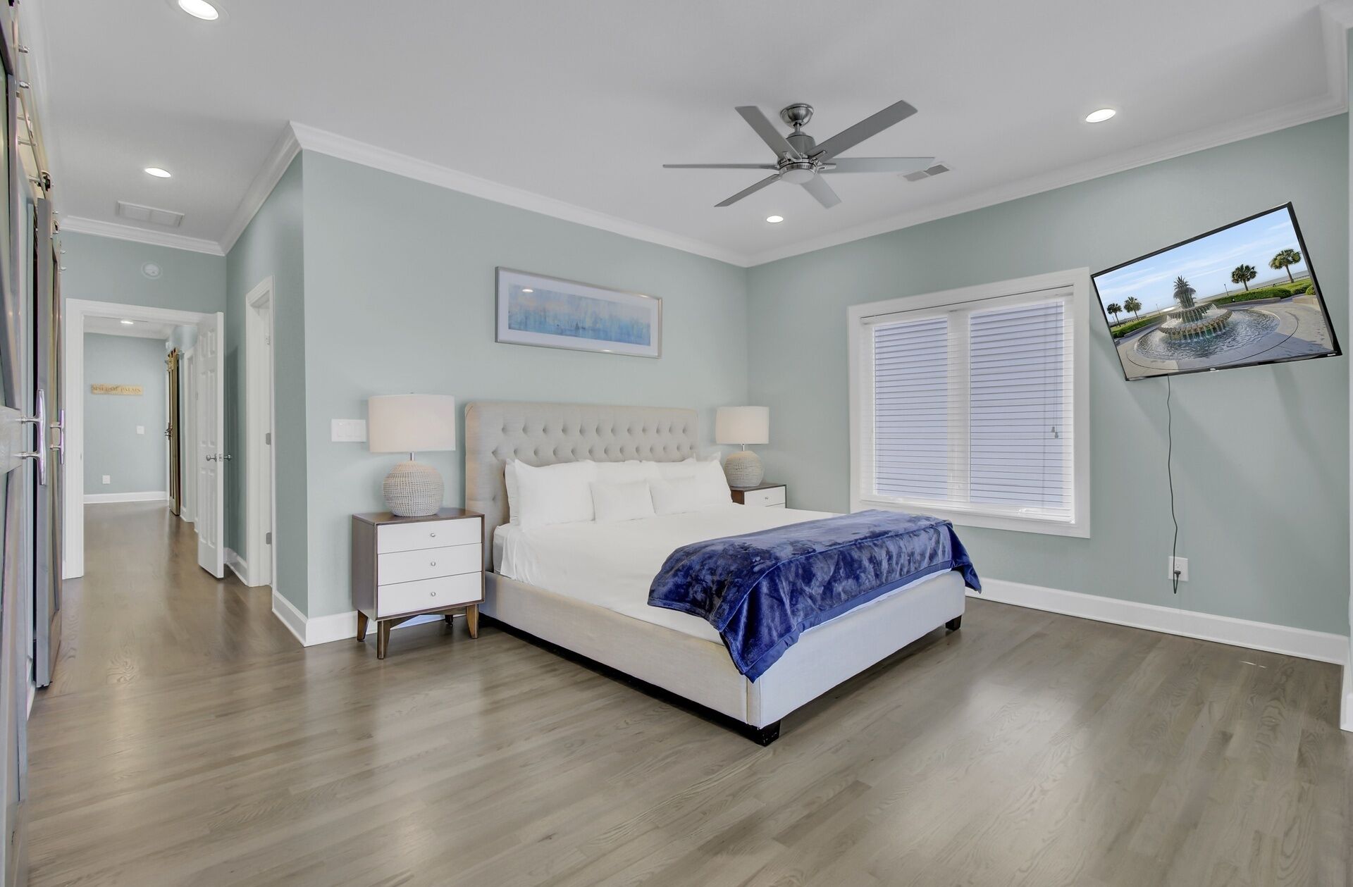 A bedroom with a bed, nightstands, a ceiling fan, a wall-mounted TV, and a window with blinds.