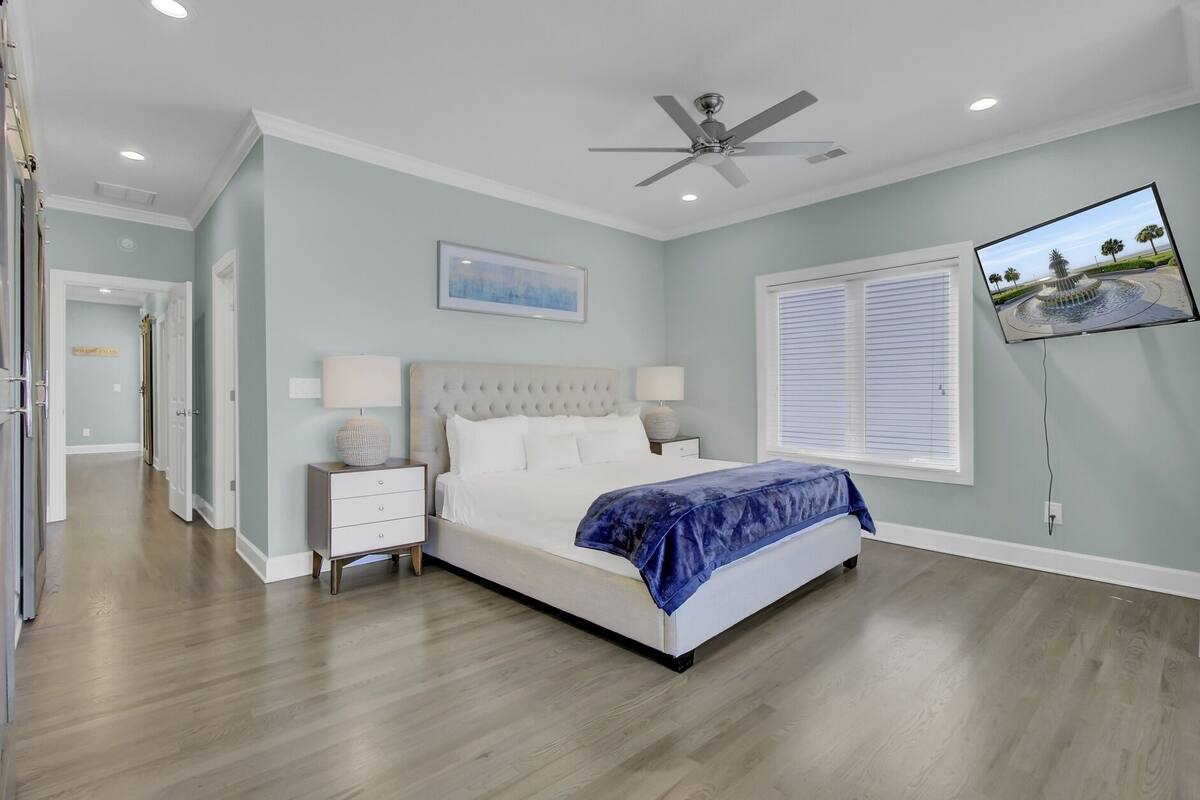 A bedroom with a bed, nightstands, a ceiling fan, a wall-mounted TV, and a window with blinds.