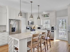 A kitchen with white cabinetry, a stainless steel refrigerator, a microwave, and a coffee maker. There are three pendant lights, a wooden island with chairs, and a view of the outdoors through a glass door. - Wild Dunes Vaction Homes 3&4 Bedrooms (Isle of Palms)