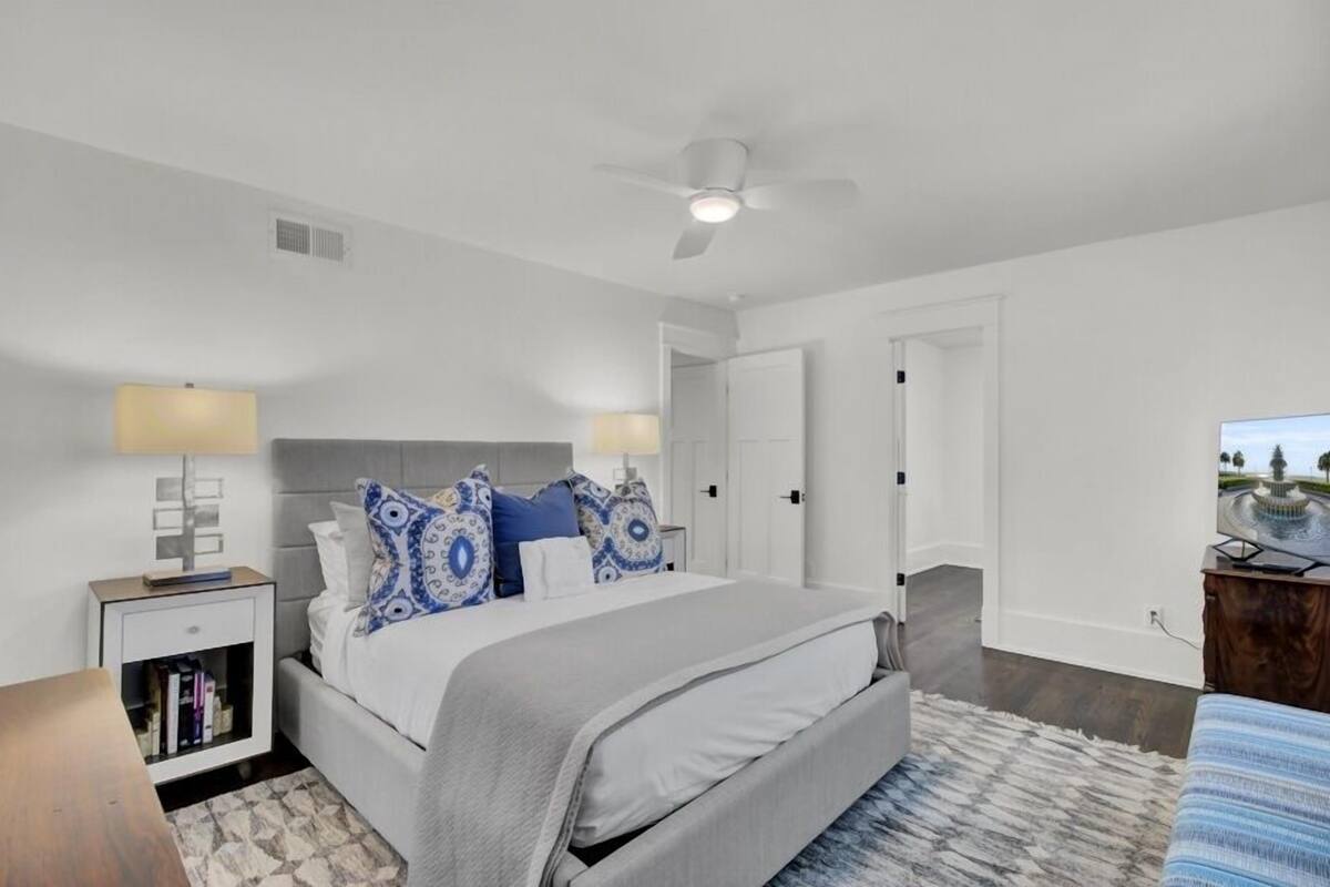 A bedroom with a bed, nightstands, a desk, a ceiling fan, and a view of a fountain.