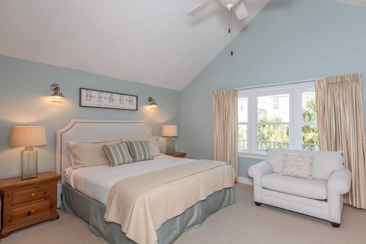 A bedroom with a bed, bedside lamps, a white chair, a window with curtains, and a ceiling fan.