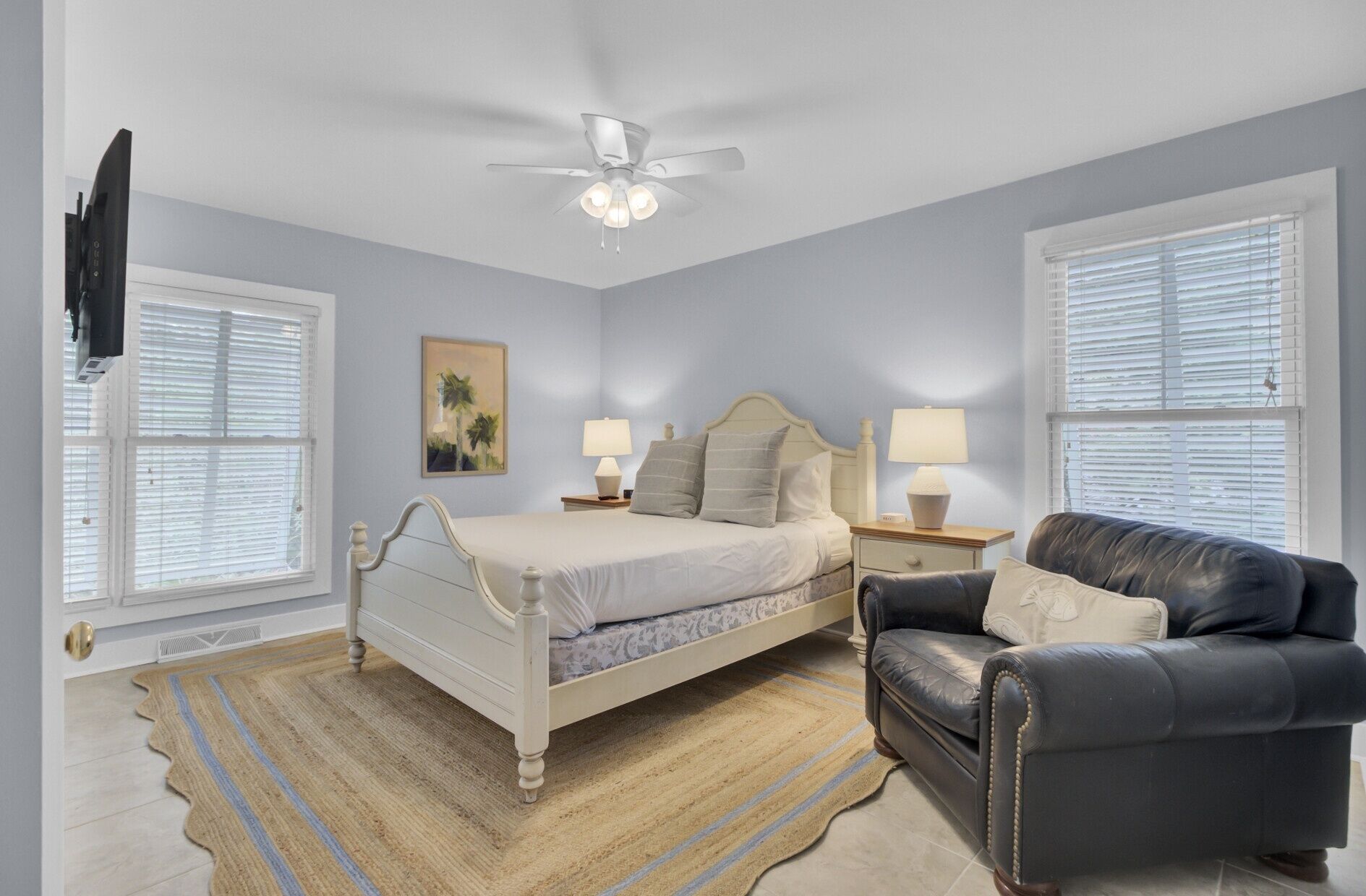 A bedroom with a bed, two bedside tables with lamps, a ceiling fan, a black leather chair, and a painting on the wall.