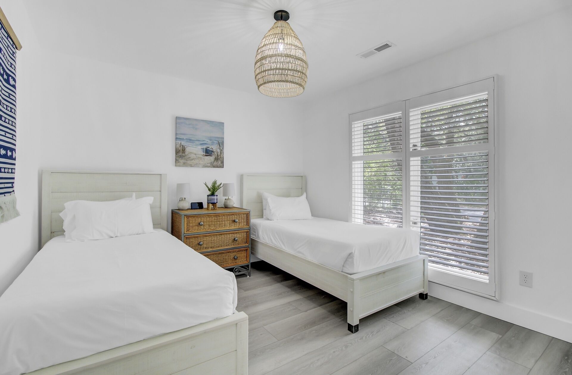 A bedroom with two beds, a nightstand, a wicker chest of drawers, a hanging light fixture, a beach-themed painting, and a window with blinds.