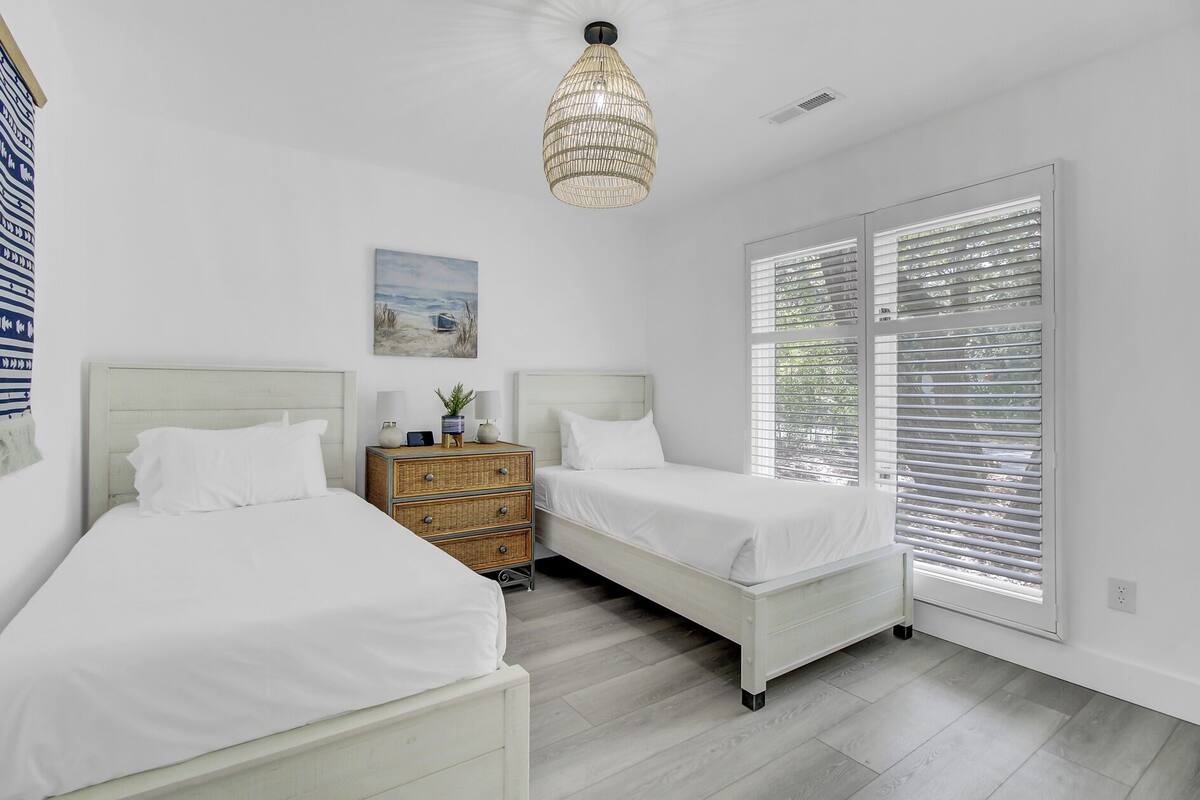A bedroom with two beds, a nightstand, a wicker chest of drawers, a hanging light fixture, and a beach-themed painting.