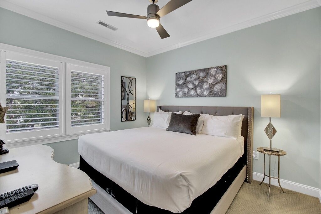 A bedroom with a bed, ceiling fan, artwork, bedside lamps, and a window with blinds.