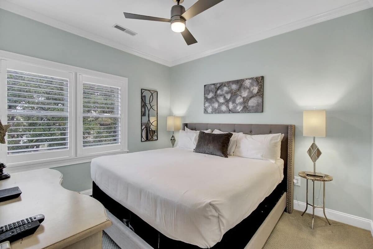 A bedroom with a bed, ceiling fan, artwork, bedside lamps, and a window with blinds.