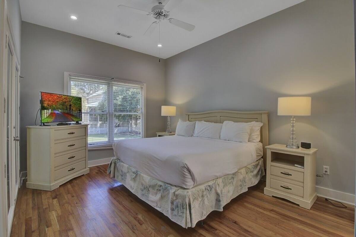 A bedroom with a bed, two bedside tables, a ceiling fan, a window with blinds, and a television on a dresser.
