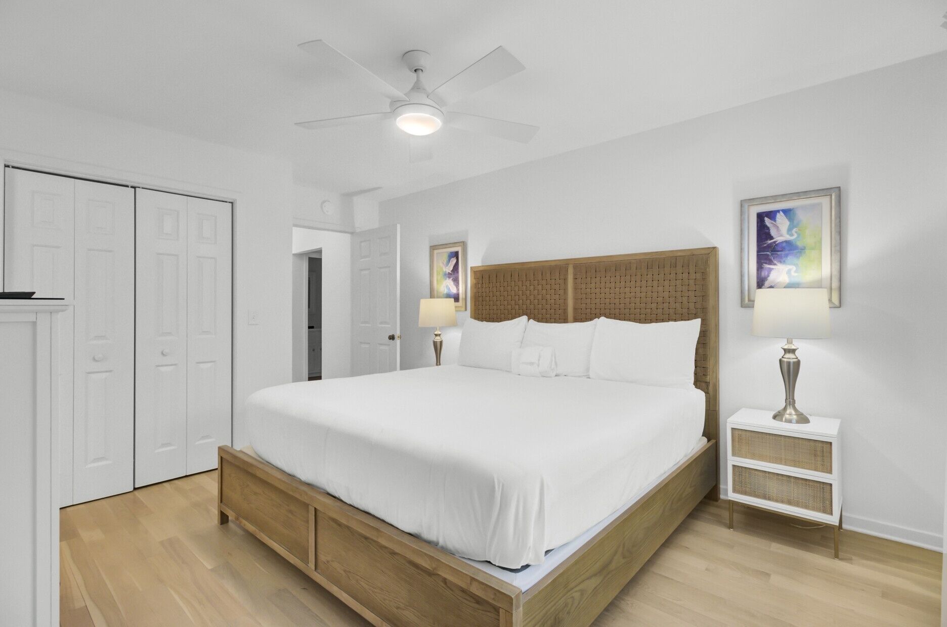 A bedroom with a wooden bed, white walls, a ceiling fan, and a nightstand with a lamp.