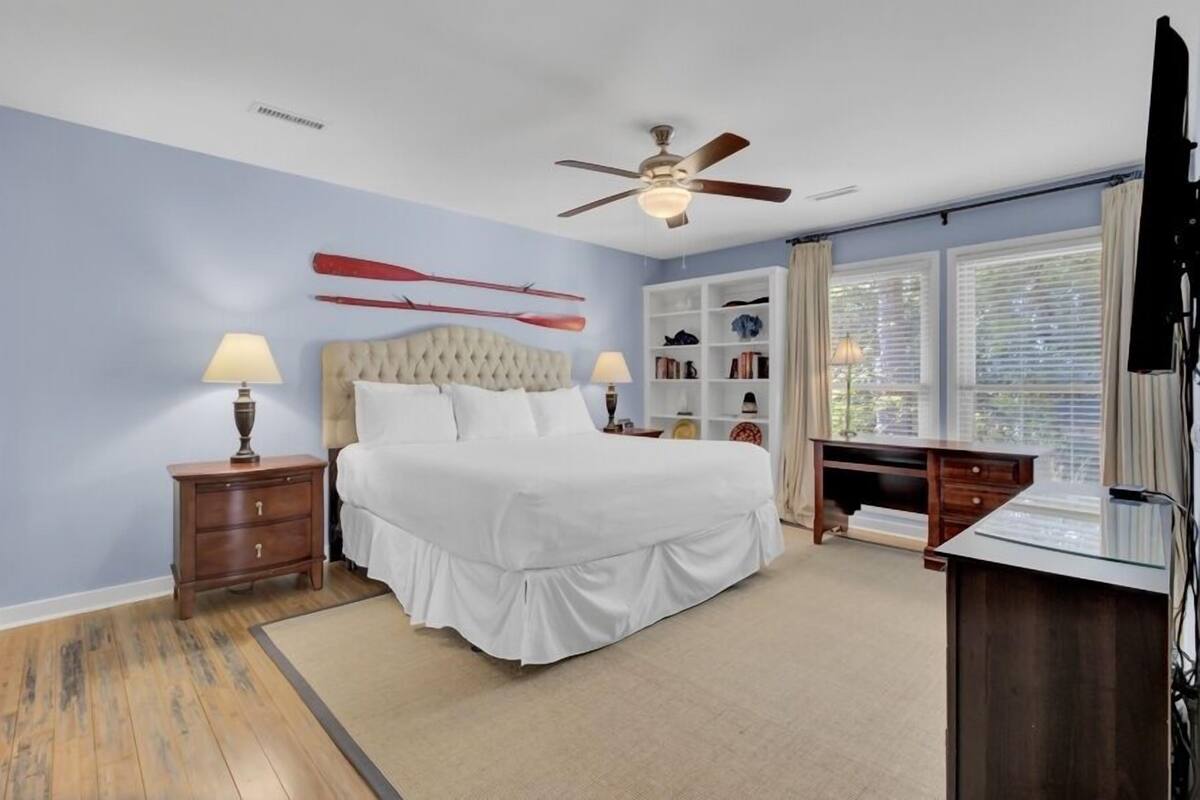 A bedroom with a bed, nightstands, a desk, a ceiling fan, and a window with blinds.