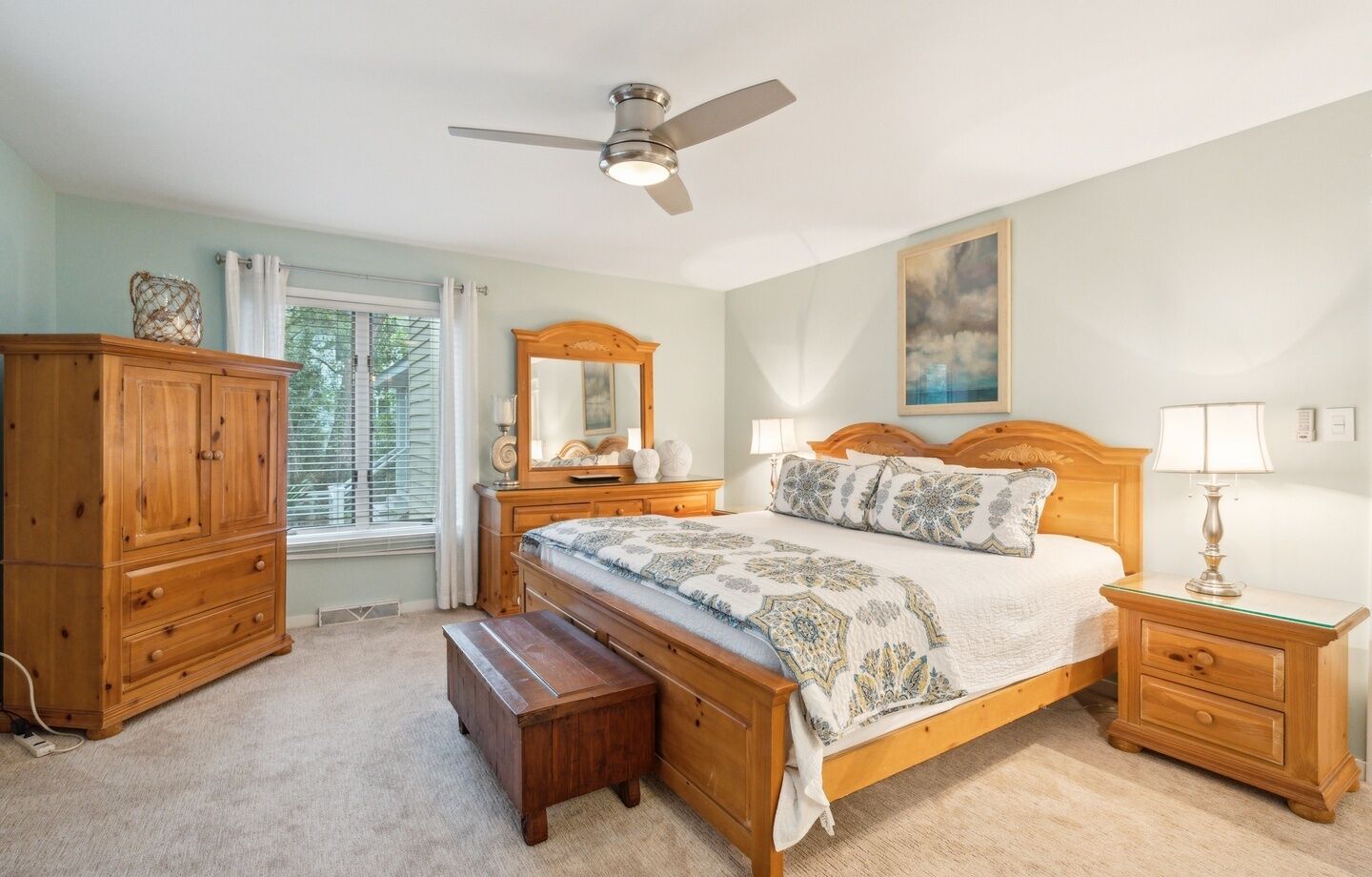 A bedroom with a bed, nightstands, a wooden dresser, a bench, a ceiling fan, and a window with curtains.