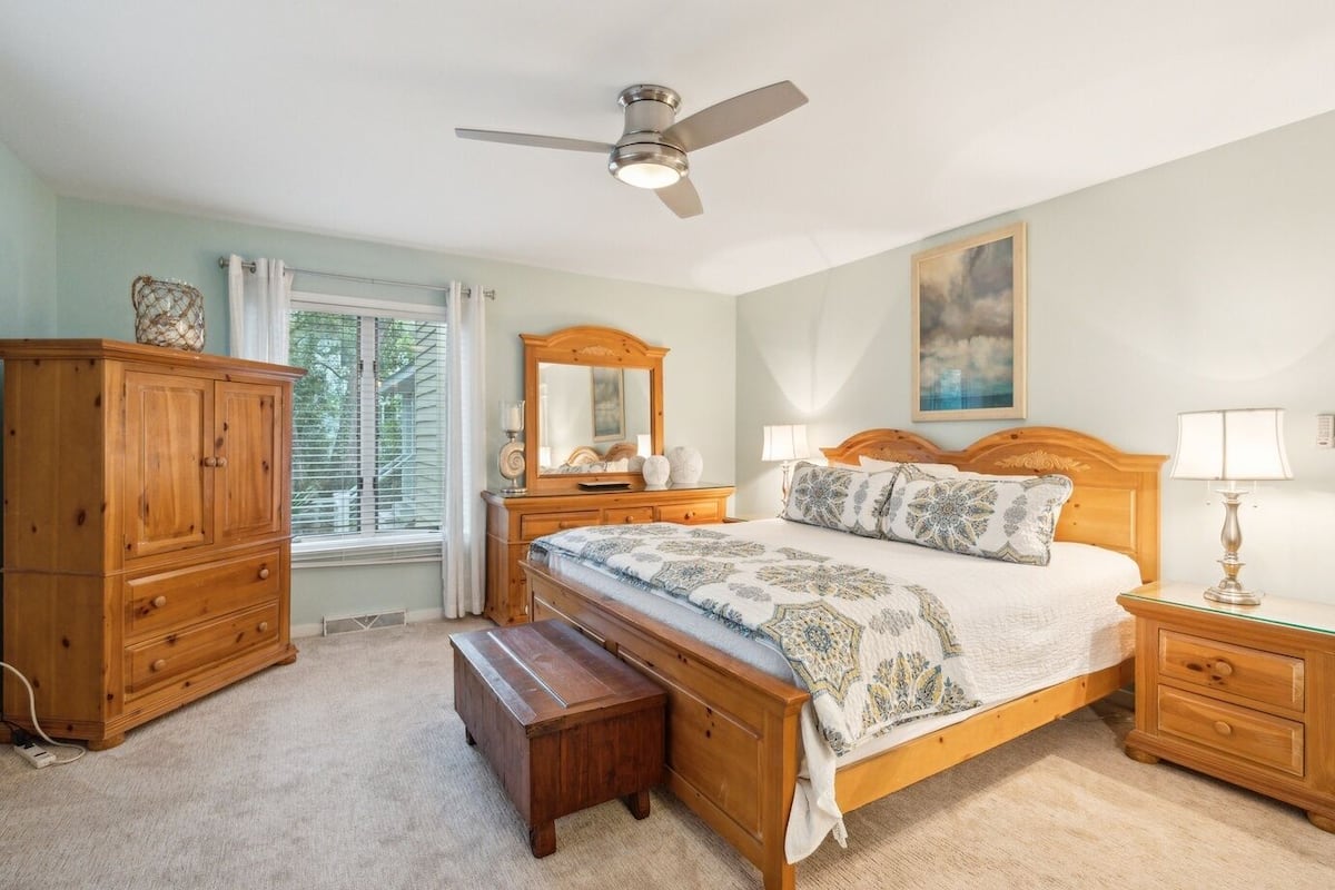 A bedroom with a bed, nightstands, a wooden dresser, a bench, a ceiling fan, and a window with curtains.