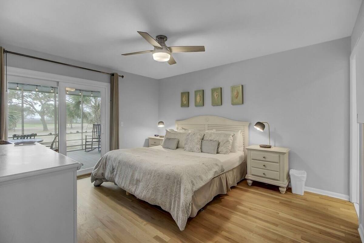 A bedroom with a bed, bedside tables, a ceiling fan, and a view through a large window.