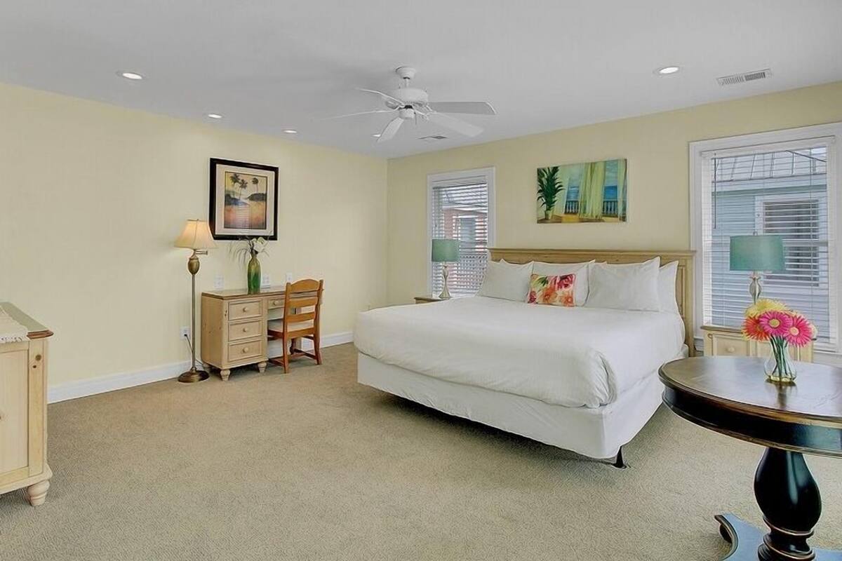 A bedroom with a bed, a desk, a chair, a ceiling fan, and a round table with a vase of flowers.